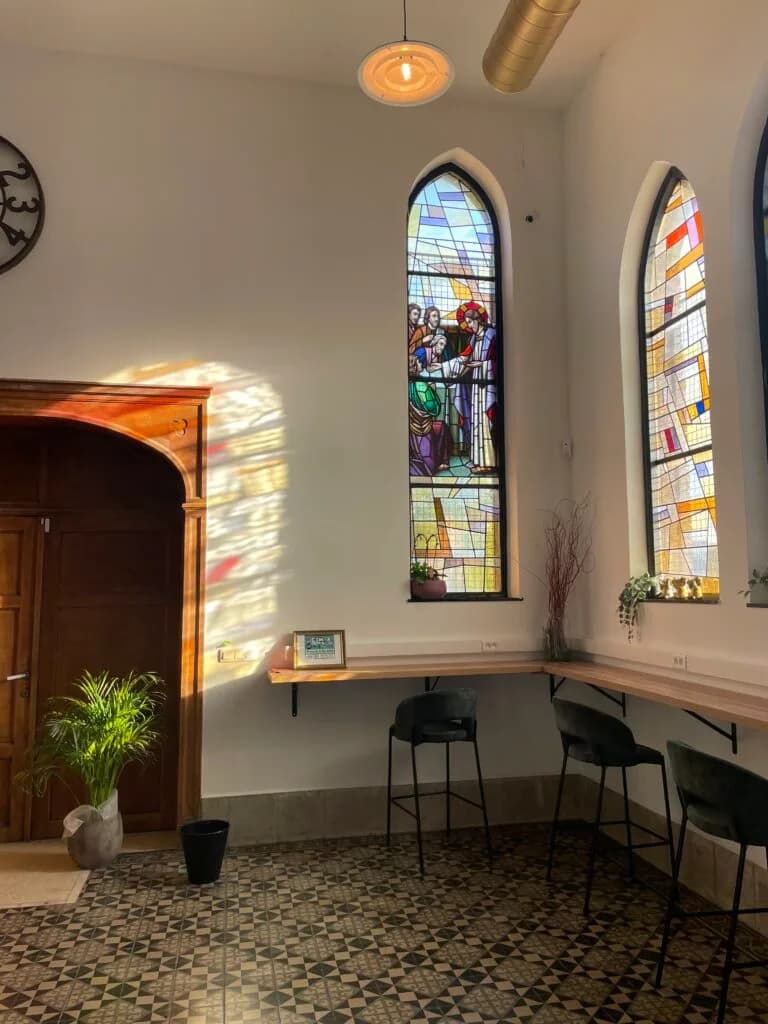 Intérieur d'un espace avec des vitraux colorés, des murs blancs, des barres de comptoir, et des sièges noirs, où une plante verte ajoute une touche de nature.